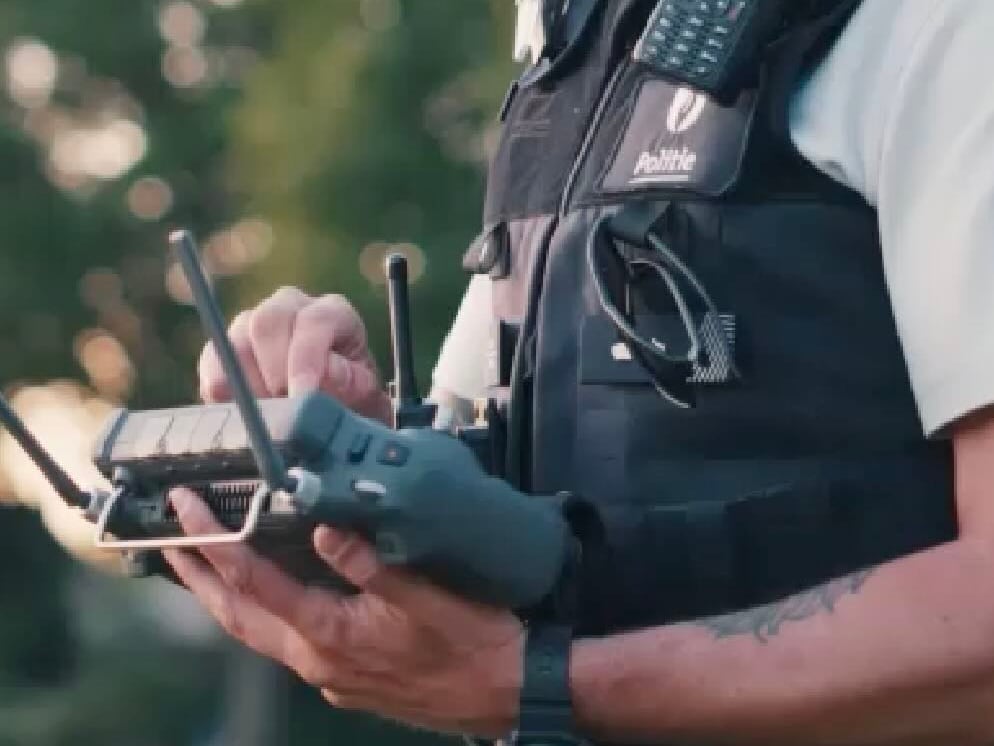 A Belgian police officer's hands on a drone controller, with a radio and body armor marked Politie