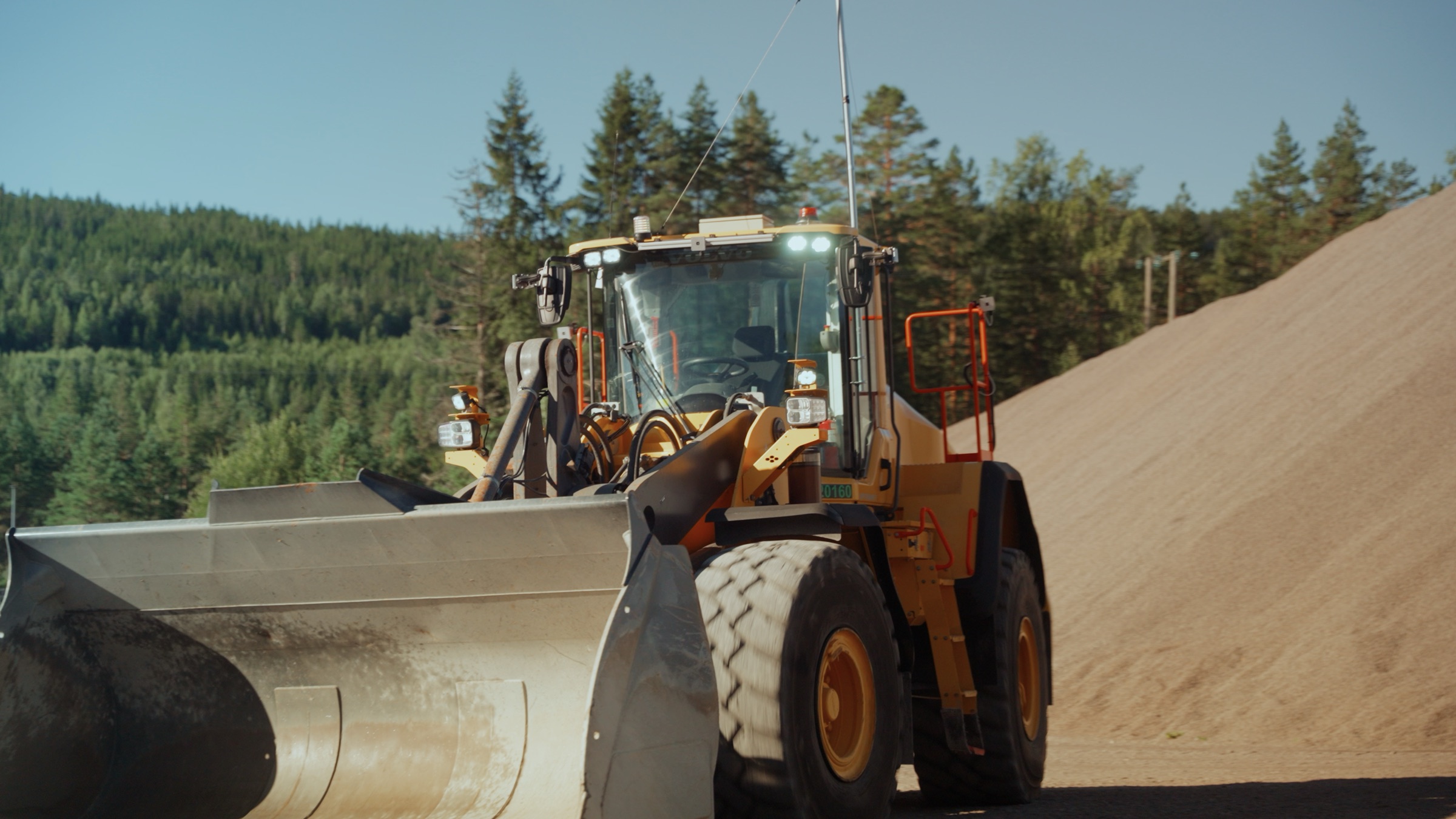 A Volvo wheel loader fitted with HIVE sensor hardware, working in the quarry at Feiring Bruk with forest and an aggregate stockpile behind it