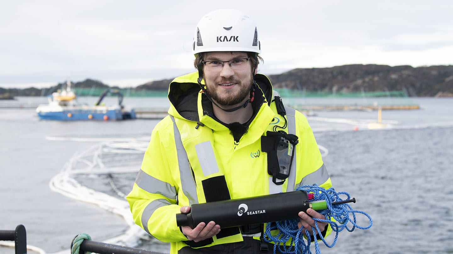 A WSense engineer in high-visibility gear holding an underwater node at a Norwegian coastal aquaculture site